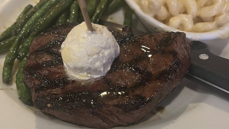 Close up of ribeye with butter with green beans and mac and cheese in background on a white plate