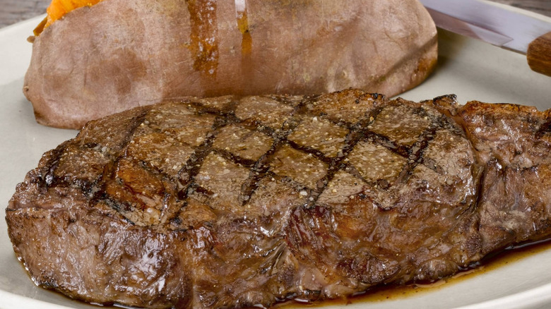 Close up of ribeye with baked sweet potato in the background on a white plate