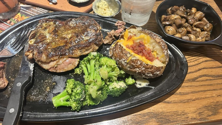 Ribeye steak with loaded baked potato, broccoli, and mushrooms on a black plate