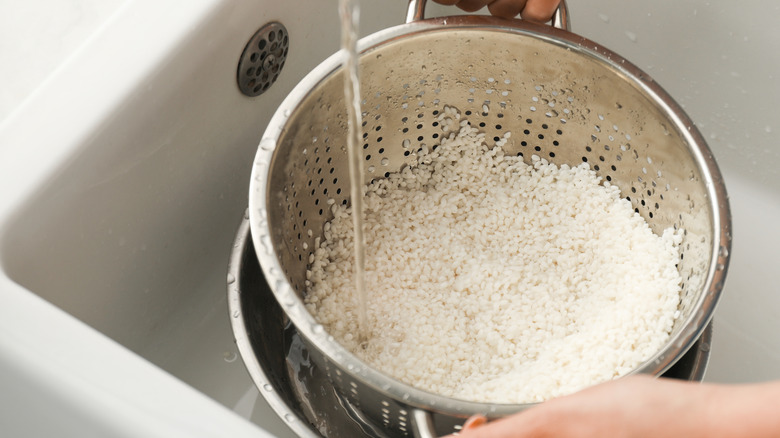 Two hands rinsing dry, uncooked white rice in a colander in the sink.