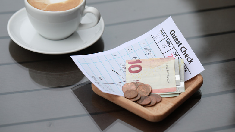 Guest check with pennies and Euro bills on small wooden tray next to cup of coffee.