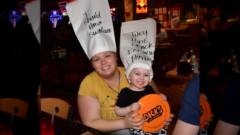 A woman holding her young child wearing hats that say "I should have swallowed" and "They hide crack in my diaper"