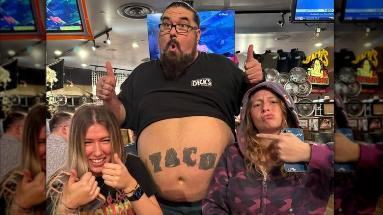 Dick's Last Resort's employee named Taco posing with his belly out next to two amused guests