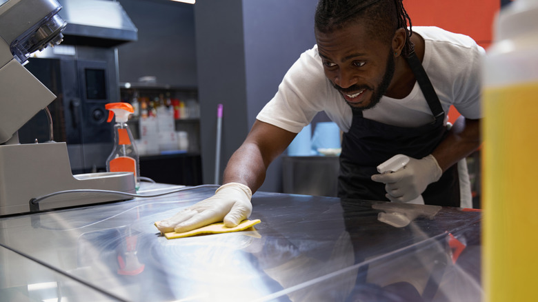 A man in an apron and gloves wiping down a counter holding cleaning supplies
