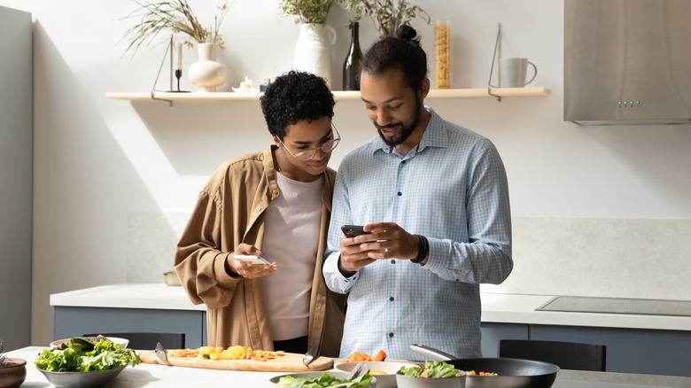 A man and a woman looking at a phone while chopping ingredients for a meal