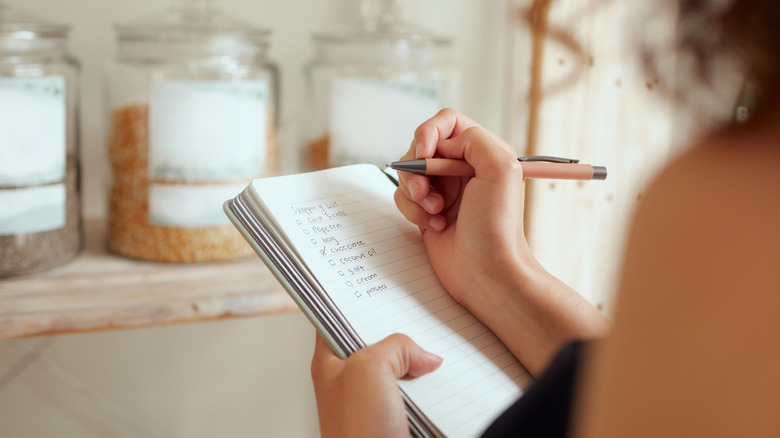 A woman writing a shopping list while looking at a shelf in her pantry