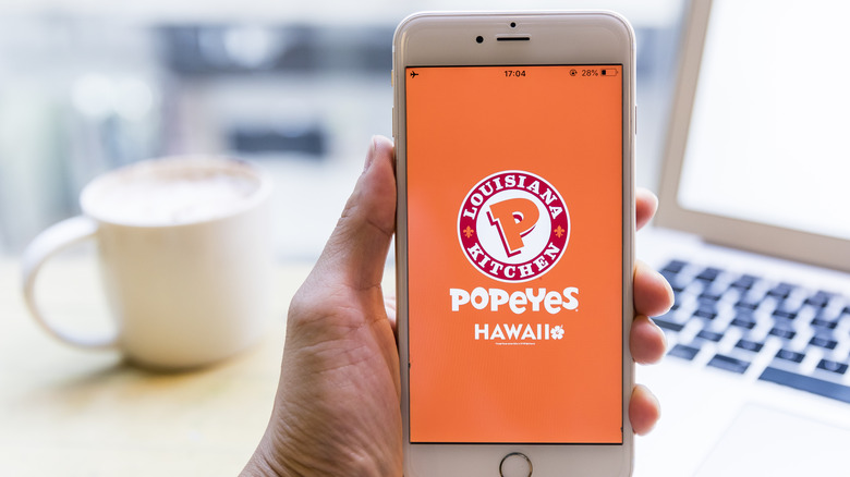 A hand holding a smartphone showing the Popeyes app logo