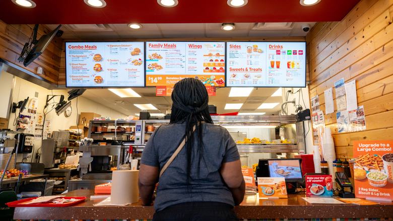 A woman stands in front of the counter at Popeyes, her back to the camera, looking up at the menu