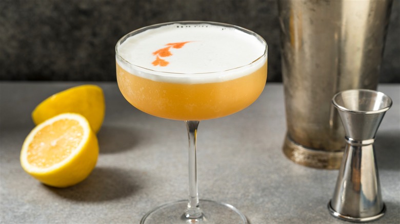A whiskey sour on a table with a shaker, a jigger, and two lemon halves.