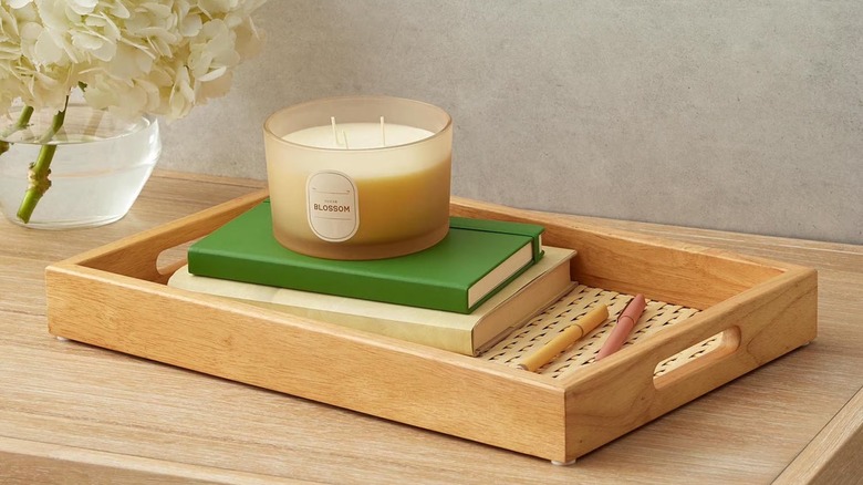 A woven serving tray holding books and a candle on a wood table.