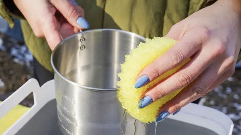 Closeup of person's hands washing a metal cup with a yellow Scrub Daddy sponge outdoors