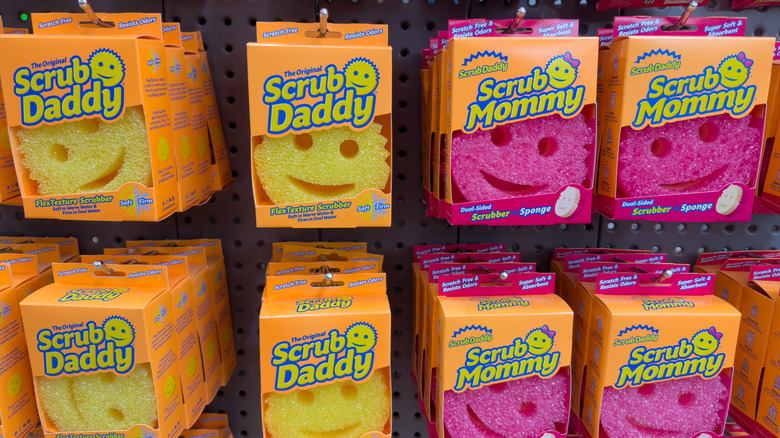 Yellow Scrub Daddy and pink Scrub Mommy sponges hanging from pegs in a store
