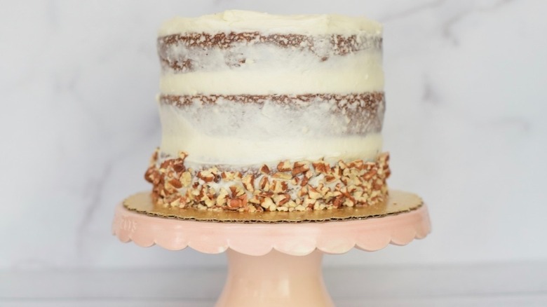 3 tiered hummingbird cake covered in frosting and chopped pecans sitting on a cake stand