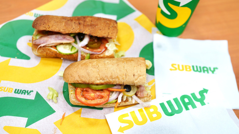 two Subway sandwiches sit on paper packaging with green and yellow logos on it