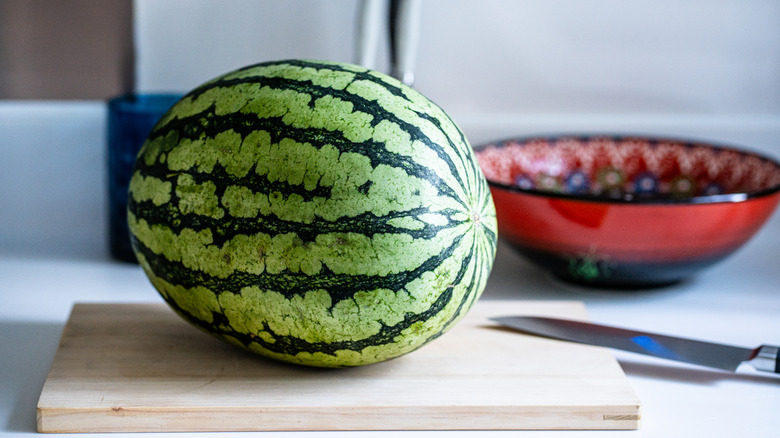 A whole watermelon sitting on a cutting board with a knife next to a red bowl