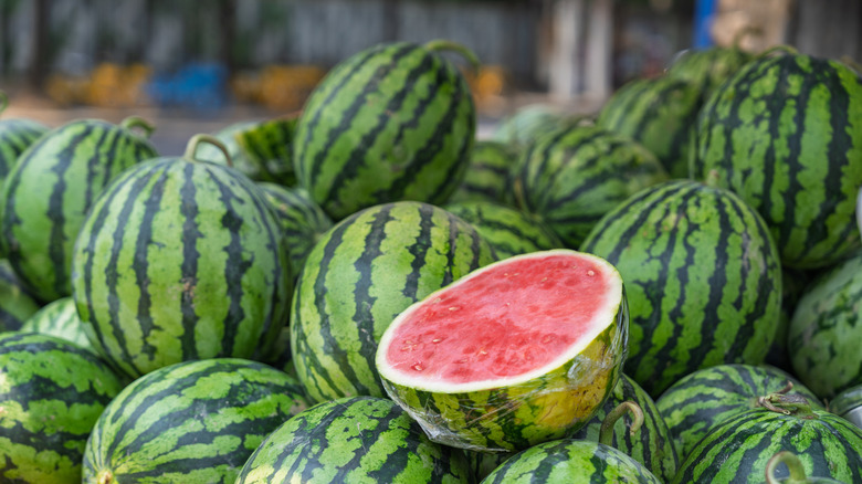 A pile of watermelons with green rinds and one cut in half with a red interior