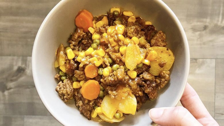 Close-up of a hand holding a bowl of shipwreck casserole: a dish with beans, ground beef, and vegetables.