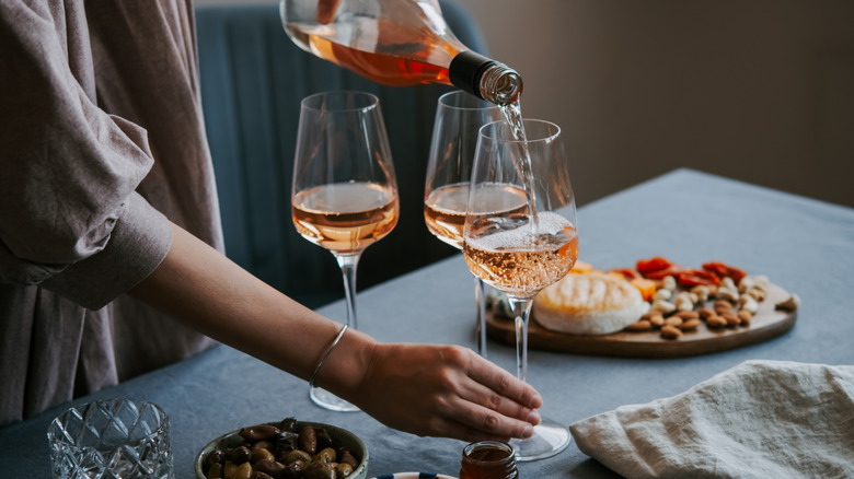 Person pouring rosé wine from bottle into three fluted glasses on table with assorted finger foods.