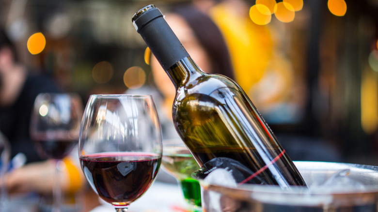 A glass of red wine on a table next to a bottle of red wine being chilled in an ice bucket