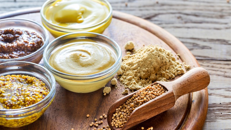 Four different kinds of mustard on a wooden board, with mustard seeds and mustard powder on display.