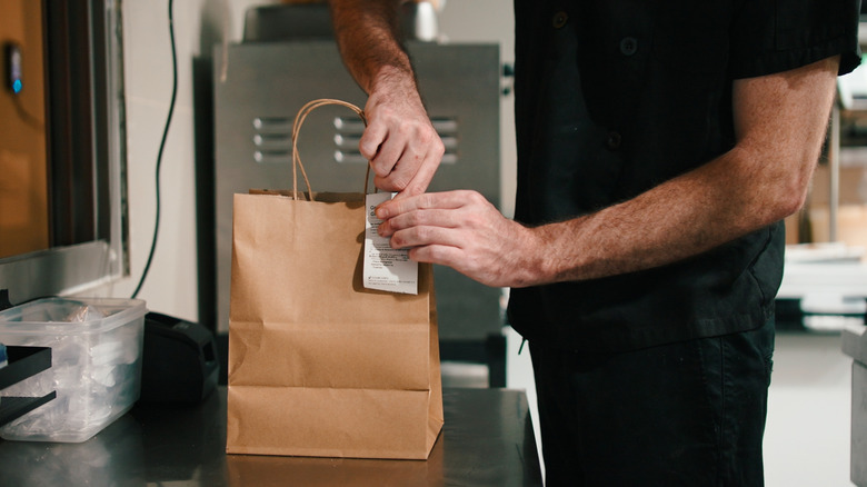 A person in black clothing prepares a brown paper bag of takeout food in a restaurant kitchen