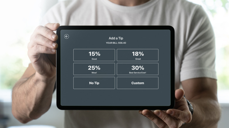 A person in a white t-shirt holds up a tablet with options to add a tip to a bill