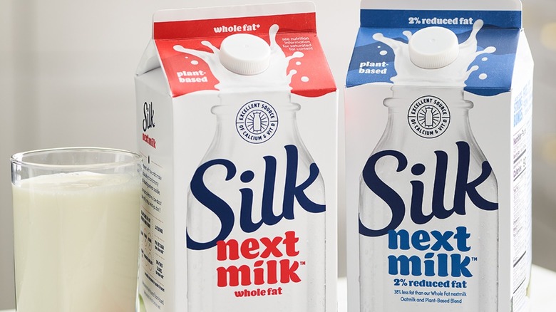 glass of milk next to two cartons of Silk NextMilk, one is whole fat, and the other is 2% reduced fat
