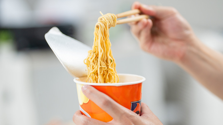 Hands pulling ramen noodles from instant cup with chopsticks.