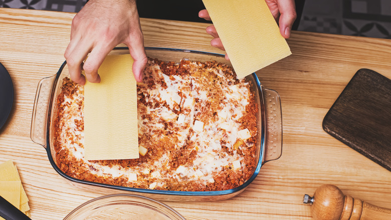 Hands assemble a lasagna, placing noodles on top of a cheesy and meaty layer in a glass baking dish.
