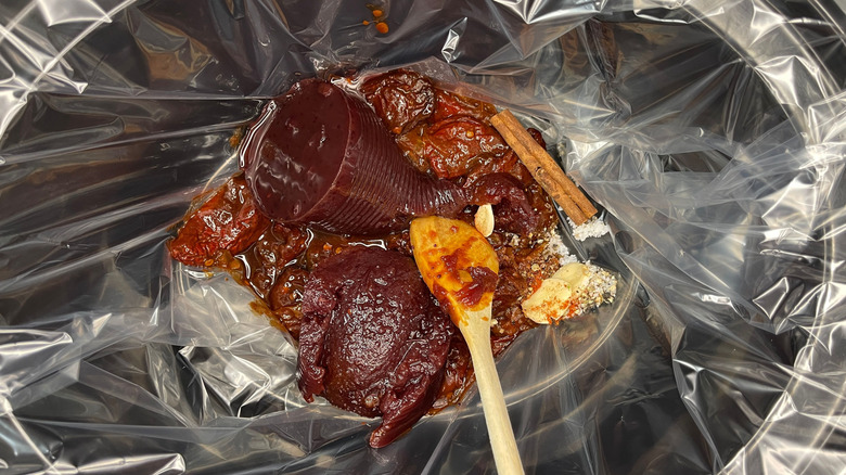 Canned cranberry sauce, spices, and other ingredients in lined slow cooker with wooden spoon
