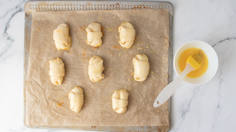 Unbaked crescent rolls on baking sheet next to bowl of egg wash