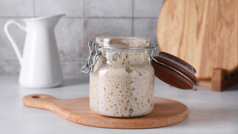 A jar of sourdough starter on wooden board with white carafe in background