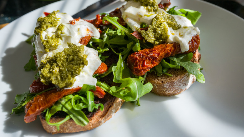 A white plate with two sourdough slices topped with greens, dried tomatoes, yogurt, and pesto