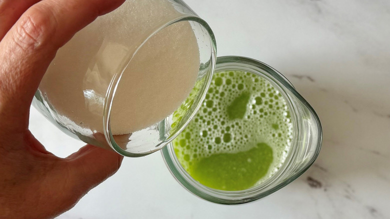 Hand pouring cup of sugar into pitcher with green liquid