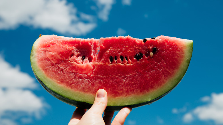 A hand holding a slice of watermelon against a blue clouded sky.