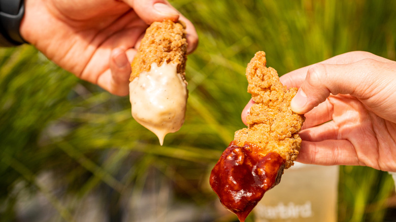 Two hands holding Starbird chicken strips, each strip dripping with Starbird signature sauce.