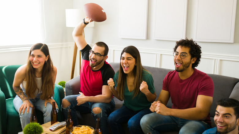 A group of friends on a couch, one a football in hand, excitedly watch football on TV.