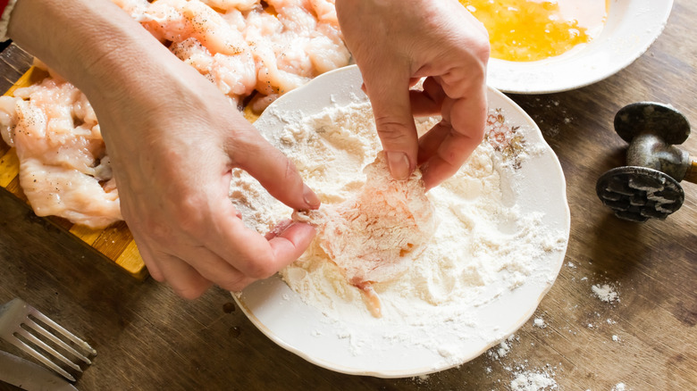 A hand dredging a chicken cutlet in flour after passing it through a beaten egg.