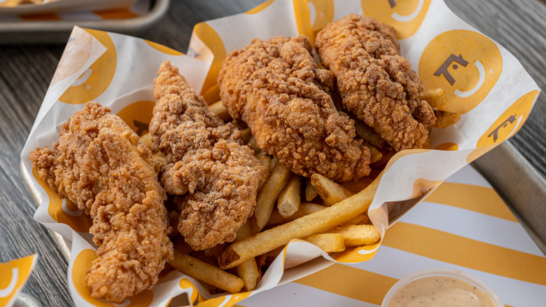 A basket of perfectly golden brown Starbird chicken tenders with fries, a ramekin of sauce below.