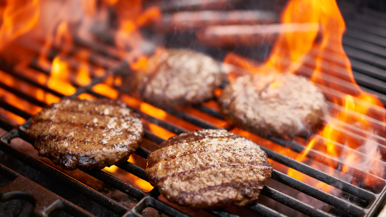 Four hamburgers cooking atop the grill grates of a flaming hot charcoal grill.