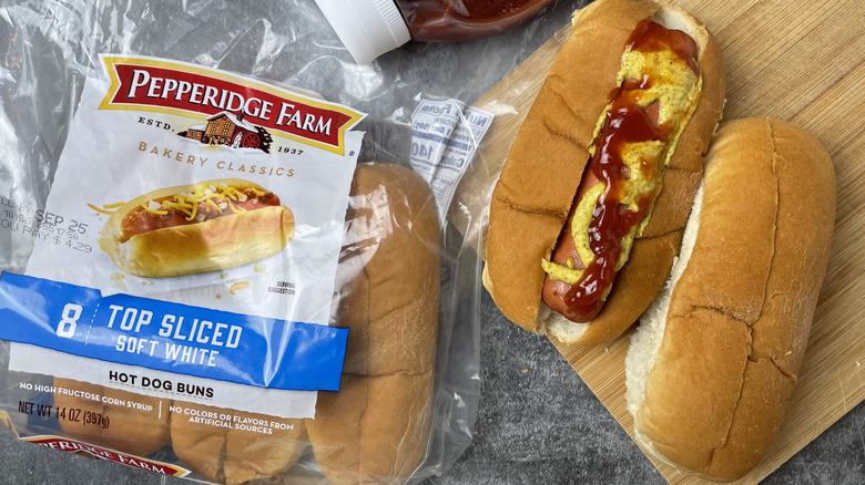 A package of Pepperidge Parm top sliced soft white hot dog buns next to a hot dog with ketchup and mustard on it