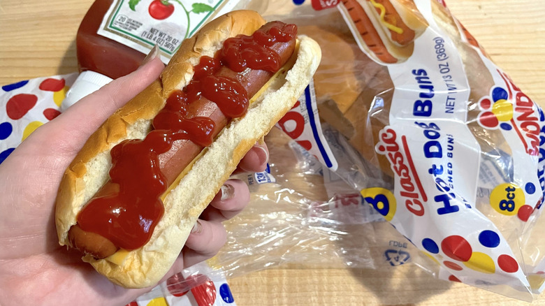 A hand holding a hot dog with ketchup on it in front of a bag on Wonder bread hot dog buns