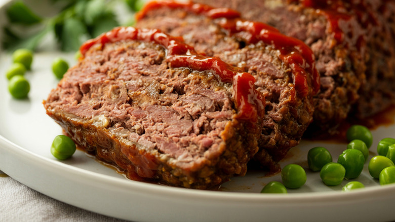 Meatloaf with a ketchup-based glaze, pieces sliced off the loaf, on a platter surrounded by green peas.