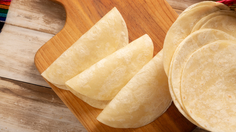 Three wheat tortillas on a cutting board over a wooden table, a pile of more tortillas to the side.