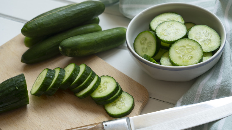 Whole and sliced green cucumbers on a wooden cutting board next to a knife