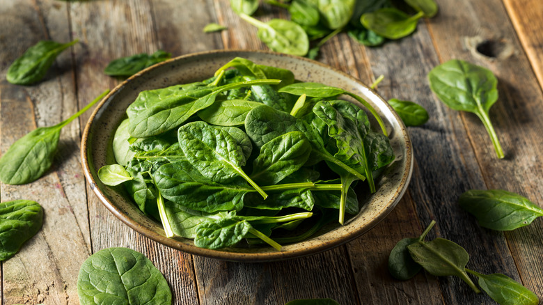 A bowl of spinach on a wooden table, surrounded by loose leaves