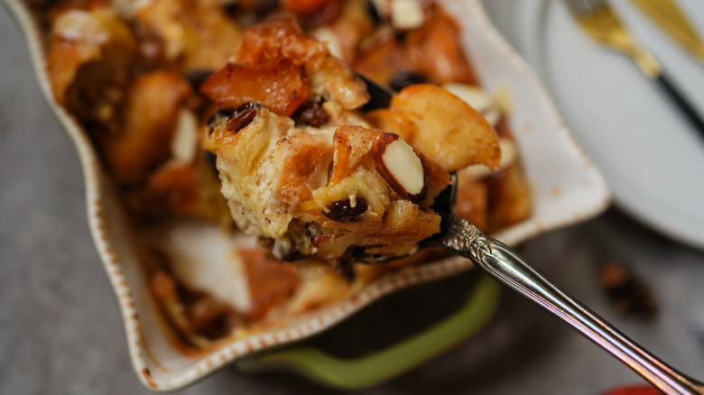 A spoon scoops bread pudding with raisins and almonds from a pan.