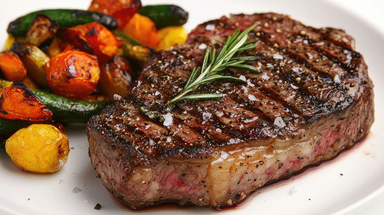 Grilled steak topped with rosemary sprig and grilled vegetables on side.