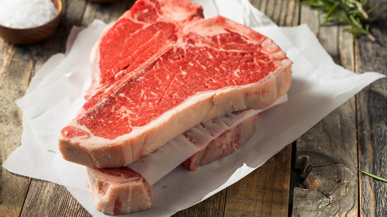 Red t-bone steaks on wax paper and a wooden background with herbs, salt, and pepper