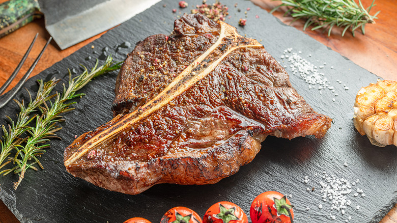 Grilled T-bone steak on a black slate with toasted herbs, roasted garlic, and charred tomatoes on the vine